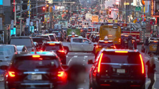 A densely packed city street filled with cars, buses, and pedestrians during dusk. Red brake lights and streetlights illuminate the scene, with towering buildings and neon signs in the background. The atmosphere is chaotic with heavy traffic and activity.