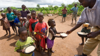 A group of children in rural Africa standing in a line to receive food from an adult. The children wear colorful but worn clothes and hold bowls as they wait, surrounded by a dry, dirt-filled landscape with green crops in the background.