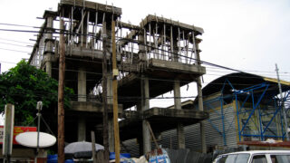 A partially constructed building with exposed concrete and wooden scaffolding. Electrical wires crisscross in front of the structure, and nearby are makeshift stalls and a satellite dish.