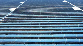 A vast array of solar panels, stretching into the distance, with rows neatly aligned. The panels reflect the blue sky, creating a sense of scale and modern energy production.