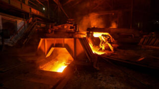 A worker, dressed in protective gear, stands near a large furnace with bright, molten metal glowing intensely. The industrial environment is dark and smoky, lit by the fiery glow of the metal.