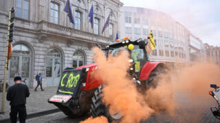 A red tractor emitting orange smoke drives through a cobblestone street in front of a government building adorned with European Union flags. The tractor is part of a protest, with signs and slogans attached to it. A few people walk nearby, seemingly unperturbed by the smoke.