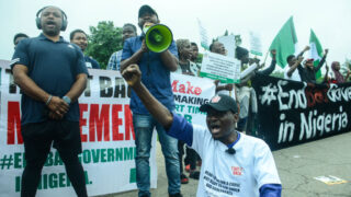 A group of protesters in Nigeria holding signs and banners, with one person speaking through a megaphone. The crowd appears passionate and unified, protesting for government reform. The background features Nigerian flags