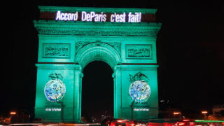 The Arc de Triomphe illuminated in green with the text "Accord De Paris c'est fait!" projected across the top, celebrating the Paris Agreement. The structure stands out brightly against the dark sky.