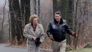 Margaret Thatcher and Ronald Reagan walking together in a wooded area during autumn. Thatcher is wearing a light-coloured overcoat, holding a black handbag, and has a concentrated expression. Reagan is dressed in a black leather jacket with a presidential seal, light trousers, and gloves, speaking animatedly with gestures. Bare trees and fallen leaves surround them.